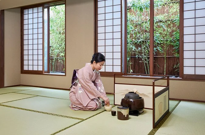 A host in a pale pink kimono kneeling on tatami mats preparing matcha beside an iron kettle, bamboo visible through the windows