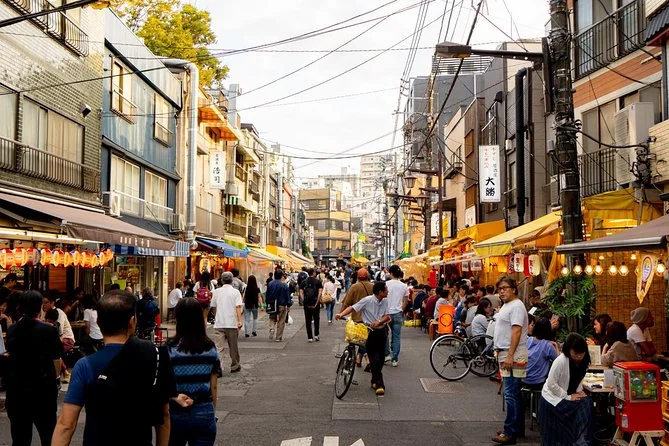 A street of open-front izakaya with lanterns and stools outside, people walking and cycling past in late afternoon light